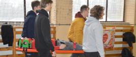 Uczymy jak ratować. Sala gimnastyczna Zespołu Szkół Zawodowych w Rawiczu. Czterech uczniów "ratowników" przenosi na noszach - typu deska "poszkodowanego" kolegę. Poszkodowany jest zabezpieczony pasami stabilizatorem głowy. 