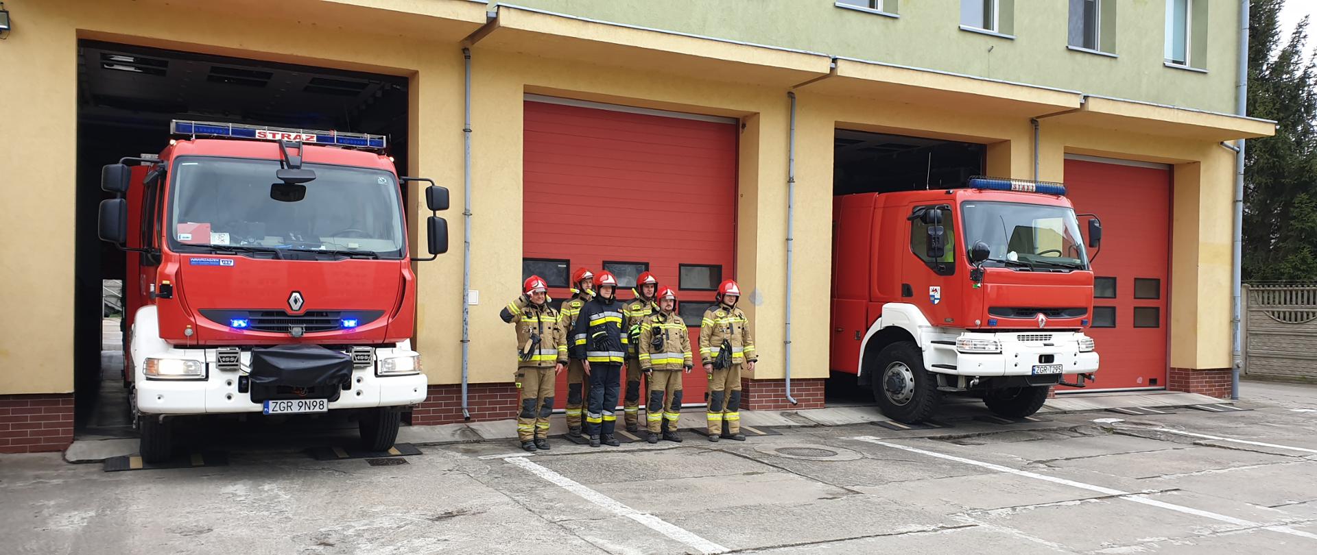 Zdjęcie zrobione na tle Komendy Powiatowej Państwowej Straży Pożarnej w Gryfinie. Na zdjęciu strażacy stoją na baczność. W tle dwa strażackie samochody stojące na sygnałach.