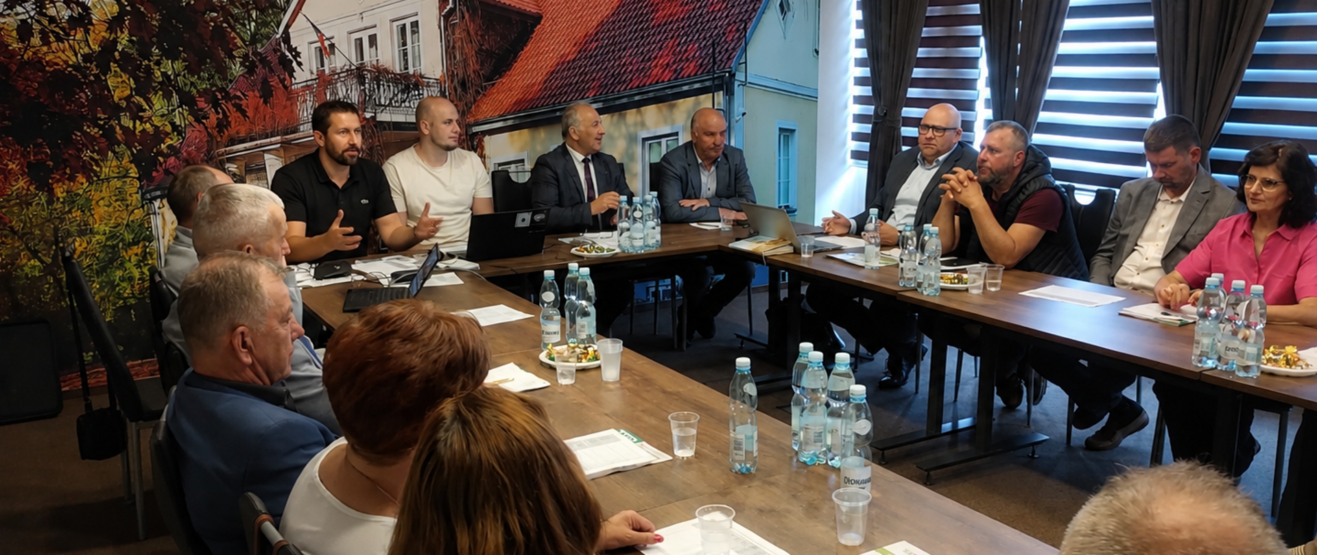 Spotkanie kilkunastu osób przy ustawionych w podkowę stołach w sali konferencyjnej. Uczestnicy prowadzą dyskusję, na stołach znajdują się dokumenty, laptopy, woda i poczęstunek. W tle widoczna jest duża grafika przedstawiająca budynek oraz okna z roletami.