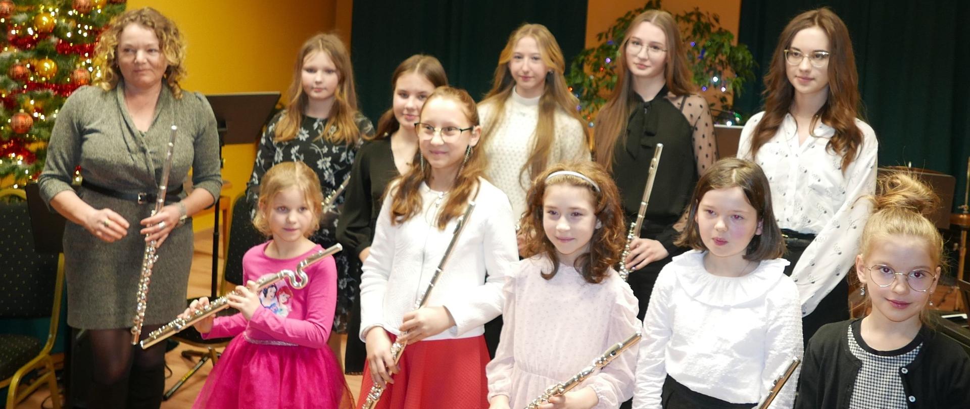 Zdjęcie grupowe uczniów klasy fletu. Wszystkie uczennice pozują do zdjęcia z fletem poprzecznym. Po lewej stronie w zielonej sukience stoi nauczycielka, która również w ręce trzyma flet. Za nauczycielką w tle stoi świątecznie ubrana choinka