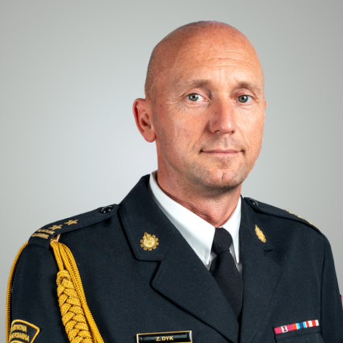 Na zdjęciu znajduje się portret mężczyzny - bryg. Zbigniewa Dyka. Mężczyzna ma owalną twarz i niebieskie oczy. Ubrany jest w ciemny mundur wyjściowy z odznaczeniami, stopniami i naszywką na ramieniu z napisem „PAŃSTWOWA STRAŻ POŻARNA”.