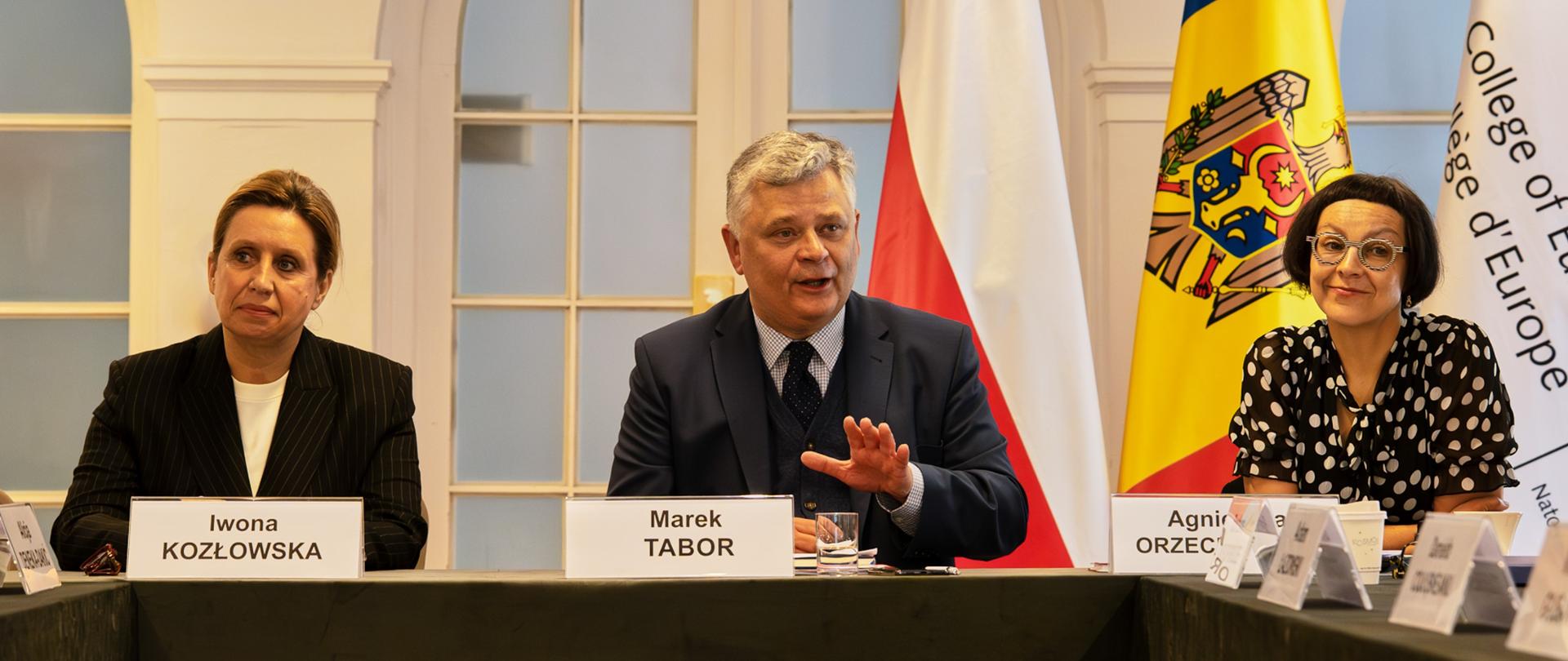 Dwie kobiety i mężczyzna siedzą przy stole konferencyjnym, przed sobą mają tabliczki z nazwiskami: Iwona Kozłowska, Marek Tabor i Agnieszka Orzechowska