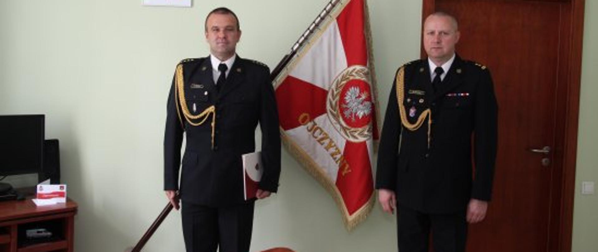 Dwóch strażaków stojących na baczność w mundurach wyjściowych ze złotym sznurem, nw tle stoi brązowe biurko, sztandar oraz godło polskie