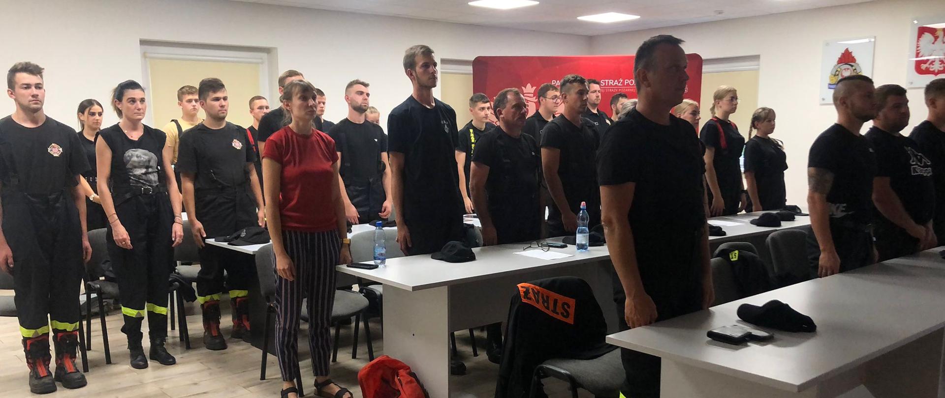 Grupa mężczyzn i kobiet stoi na baczność na oświetlonej sali, stoją przy białych stolikach, część z nich ubrana jest w mundury koszarowe straży pożarnej