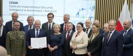 Grupowe zdjęcie uczestników uroczystości podpisania porozumienia dotyczącego Centrum Implementacji Sztucznej Inteligencji w Automatyce i Robotyce (CESAR). W centrum widoczny jest otwarty dokument prezentowany uczestnikom wydarzenia.