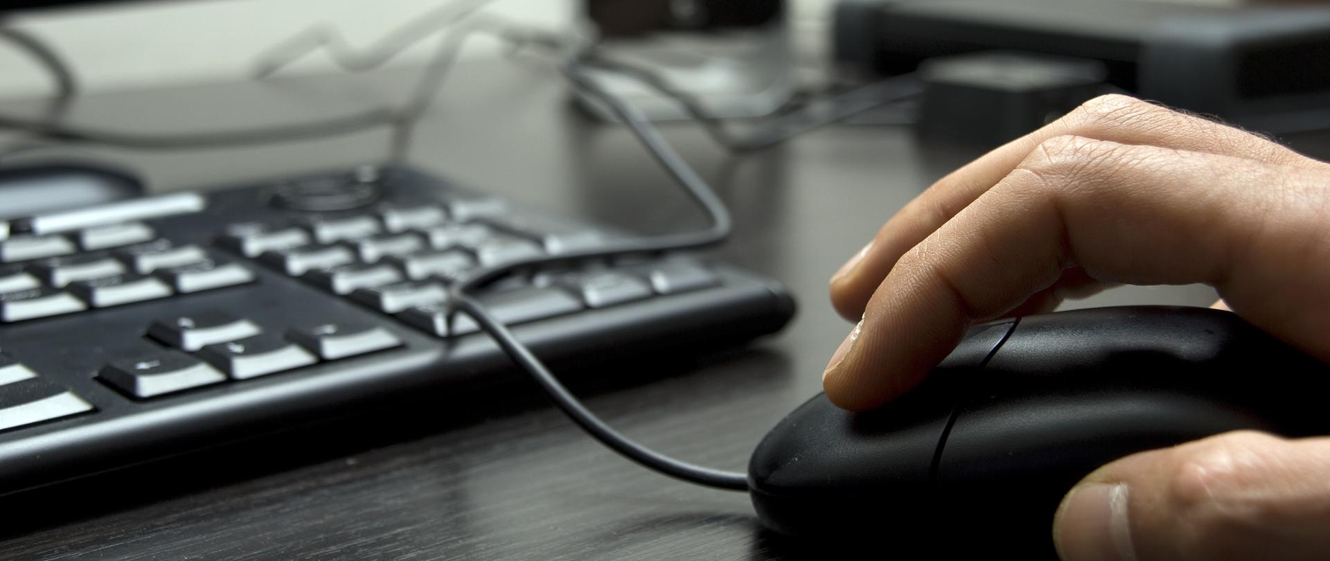 Dłoń trzymająca mysz, w tle klawiatura.