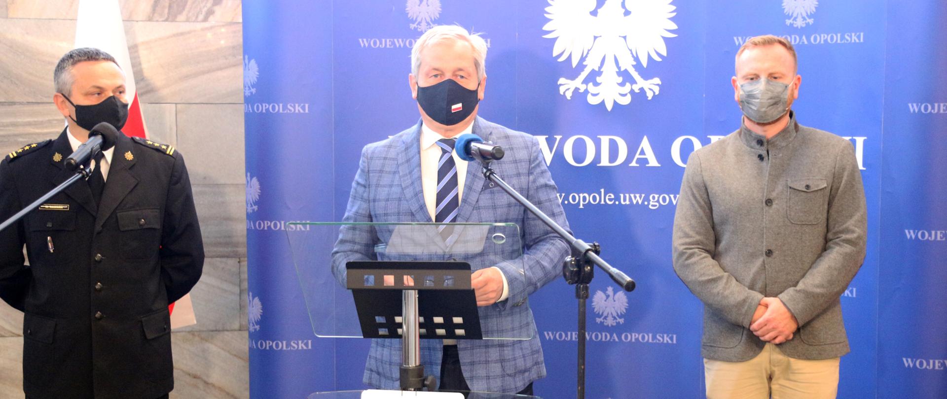 Zdjęcie z konferencji prasowej wojewody opolskiego. Na pierwszym planie, na tle granatowej ścianki z napisem Wojewoda Opolski, stoi Sławomir Kłosowski, wojewoda. Przemawia on stojąc za szklanej mównicy. Przed nim na podłodze ulokowano stojak z mikrofonem. Po prawej stronie kadru stoi z-ca kierownika Centrum Powiadamiania Ratunkowego Ditmar Kurzał, ubrany w jasne spodnie i szarą marynarkę. Spogląda przed siebie. Po lewej stronie kadru widać stojącego na tle biało-czerwonej flagi RP z-cę komendanta straży pożarnej. Meżczyzna spogląda w kierunku wojewody. Ma krotkie, ciemne włosy oraz maseczkę ochronną na twarzy. 