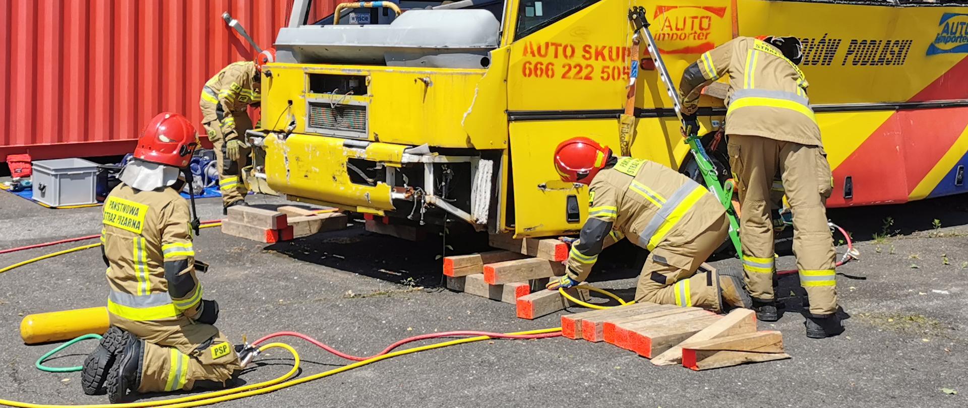 sześciu strażaków ubranych w ubrania specjalne pracuje przy raku autobusu, w tle czerwone kontenery
