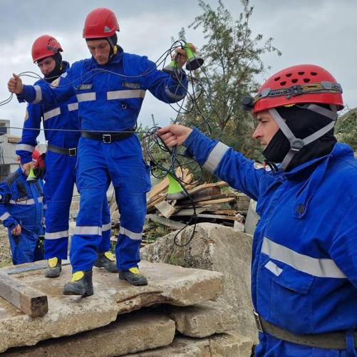 Grupa mężczyzn w niebieskich kombinezonach i kaskach stoi wokół i na płytach cementowych i rozciąga kable podłączone do sprzętu ratowniczego