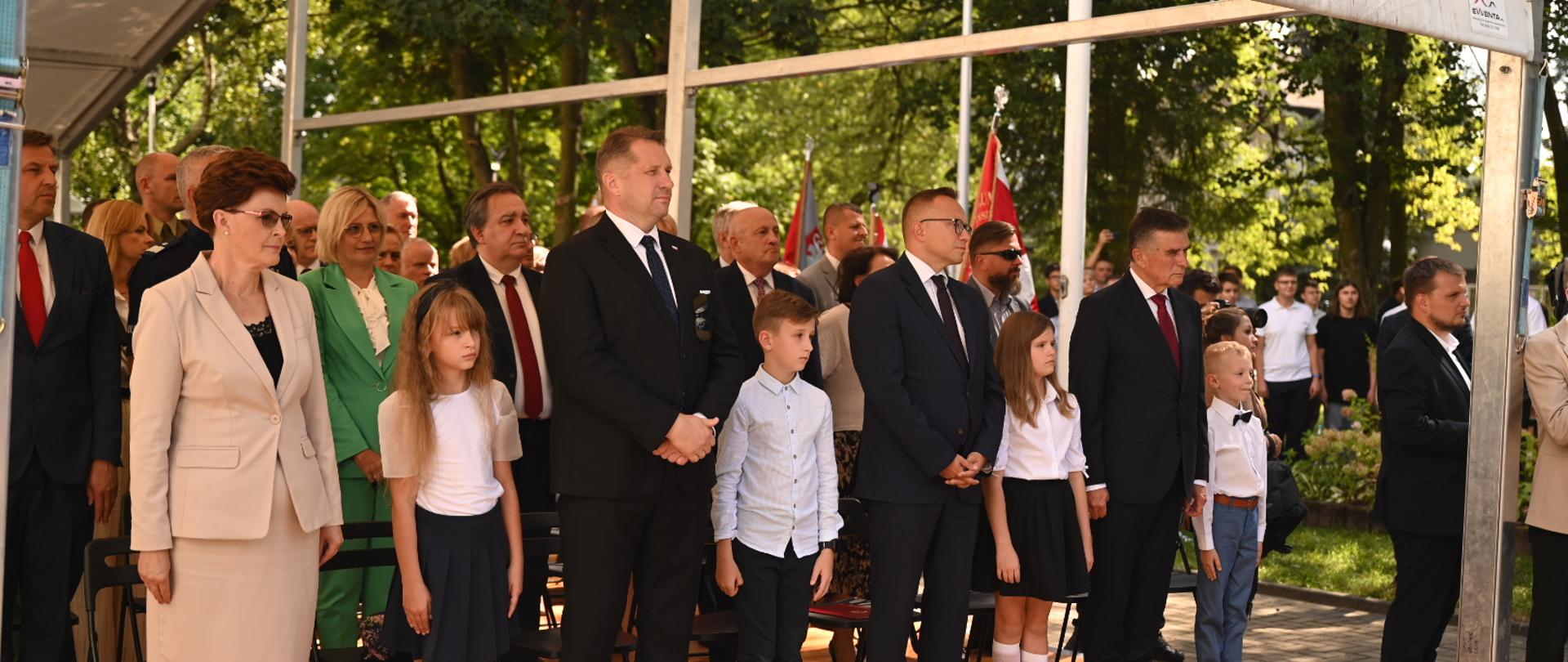 Pod namiotowym daszkiem stoi minister Czarnek, kilka innych osób i galowo ubrane dzieci, za nimi drzewa.