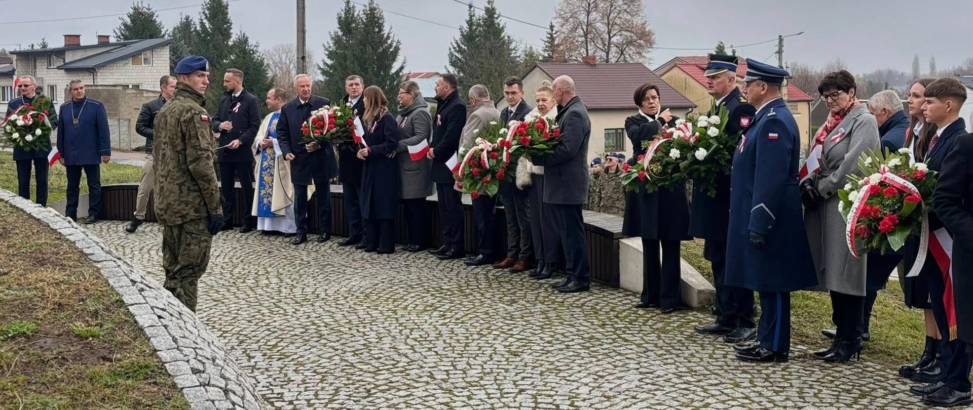 Zdjęcie przedstawia uroczystość patriotyczną lub rocznicową odbywającą się na brukowanym placu pod pochmurnym niebem.
W centralnej części kadru widoczna jest grupa około dwudziestu osób stojących w rzędzie. Większość z nich ubrana jest w eleganckie, ciemne płaszcze i trzyma duże, okrągłe wieńce lub wiązanki kwiatów w barwach narodowych (biało-czerwonych, z przewagą czerwonych róż i białych elementów, przepasane szarfami).
Wśród stojących osób wyróżniają się:
1. Po lewej stronie w rzędzie: Mężczyzna w sutannie i niebieskiej kapie (duchowny) oraz mężczyzna w cywilnym stroju, trzymający wieniec.
2. Po prawej stronie w rzędzie: Dwóch mężczyzn w mundurach galowych – jeden w granatowym mundurze policyjnym z czapką, drugi w ciemnym mundurze strażackim z naszywkami.
Na pierwszym planie, z lewej strony, stoi samotnie tyłem do aparatu żołnierz w mundurze polowym, odwrócony w stronę grupy.