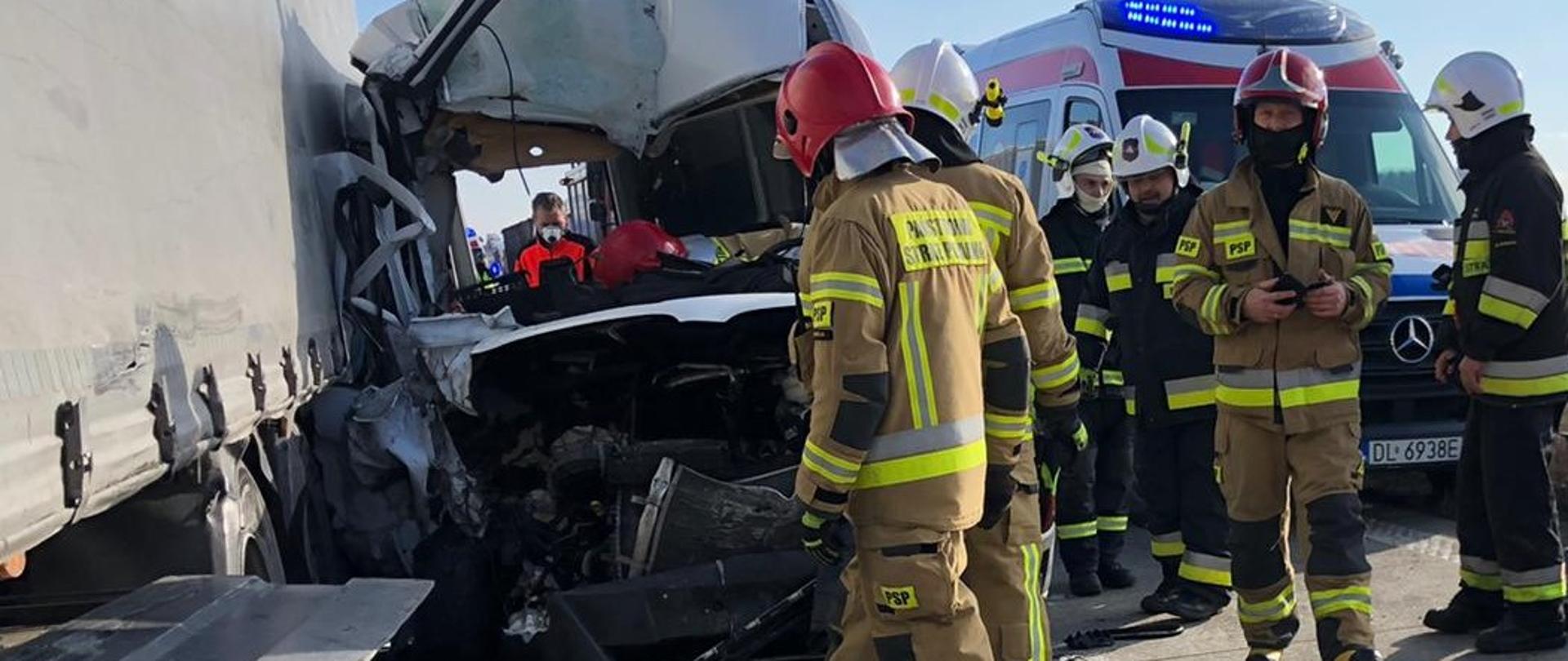 uszkodzony samochód (bus) w wyniku wypadku stojący obok naczepy samochodu ciężarowego, obok pojazdów stoją strażacy, w tle widoczny samochód pogotowia ratunkowego