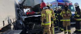 uszkodzony samochód (bus) w wyniku wypadku stojący obok naczepy samochodu ciężarowego, obok pojazdów stoją strażacy, w tle widoczny samochód pogotowia ratunkowego