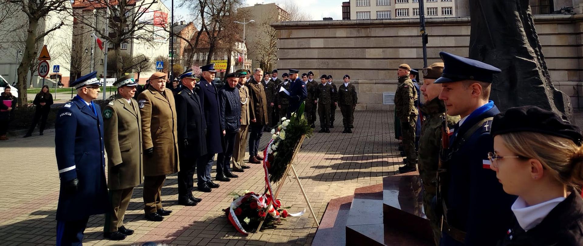 Na zdjęciu widać oficjalną uroczystość odbywającą się na zewnątrz, pod dużym pomnikiem papieża św. Jana Pawła II w Katowicach na placu katedralnym. Przed pomnikiem ustawiono wieńce z biało-czerwonymi kwiatami.
Na pierwszym planie stoi grupa szefów służb mundurowych — wojskowych, policjantów, strażaków, przedstawicieli straży granicznej. Wszyscy stoją na baczność. Za nimi widoczna jest druga linia żołnierzy i uczniów w mundurach. Obok nich stoją też osoby cywilne.
W tle widać miejską zabudowę, niskie bloki i kamienice oraz kilka drzew jeszcze bez liści. Pogoda jest słoneczna, niebo lekko zachmurzone.
