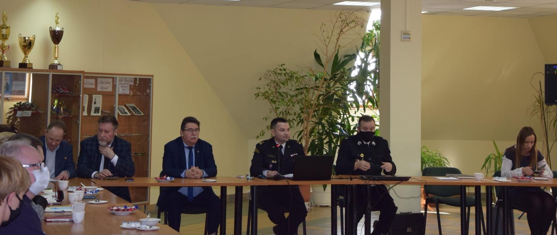 Przy stołach ustawionych na kształt przypominający podkowę, siedzą kobiety i mężczyźni. W tle widać gablotę z pucharami i dyplomami oraz kwiaty doniczkowe.