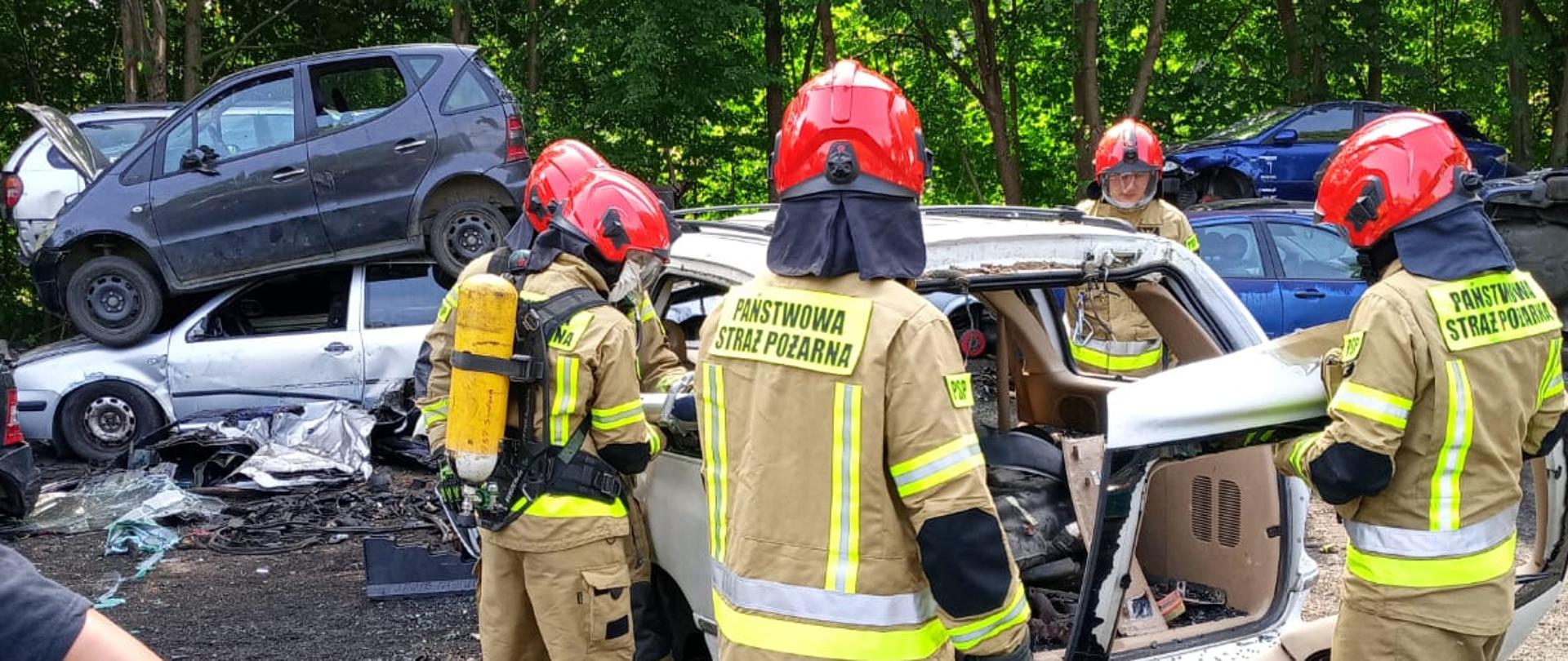 Zdjęcie przedstawia grupę strażaków podczas szkolenia podstawowego. Strażacy ćwiczą cięcie, rozpieranie elementów pojazdów. Zajęcia z tematyki: metody uwalniania osób poszkodowanych z samochodów osobowych, ciężarowych oraz autobusów – zajęcia przeprowadzane w punkcie kasacji pojazdów Eco-Auto-System w Rąbinie.