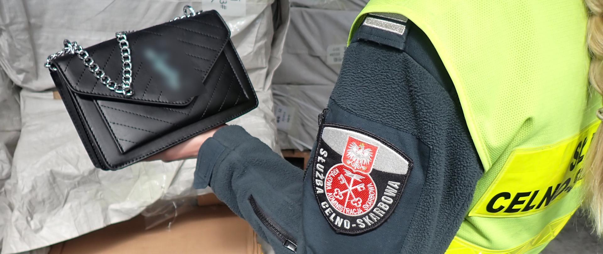 Osoba w kamizelce „Służba Celno‑Skarbowa” trzyma czarną pikowaną torebkę z łańcuszkiem na tle zapakowanych worków.