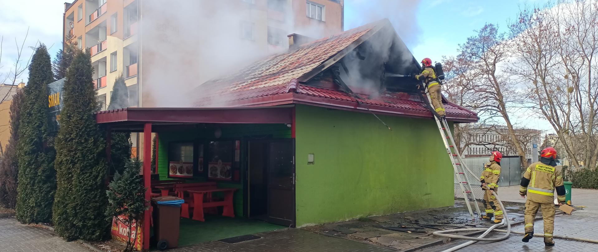 Budynek, w którym jest pożar. Na drabinie stoi strażak i gasi. Obok drabiny stoi 2 strażaków.