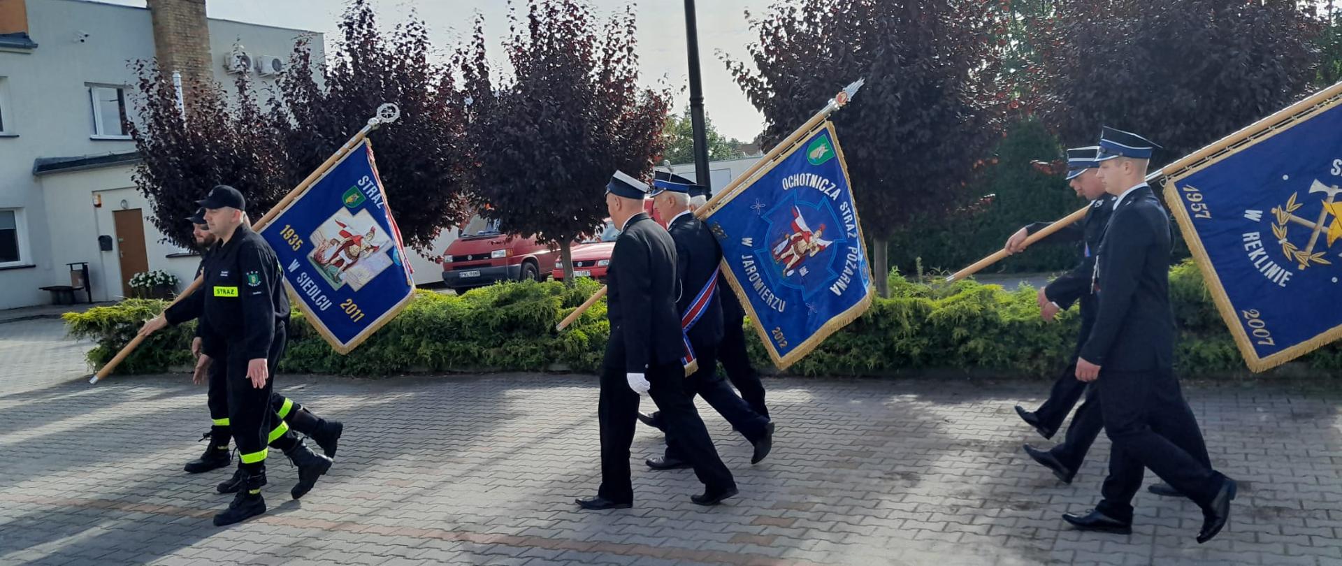 Strażacy maszerujący w trójkach ze sztandarami