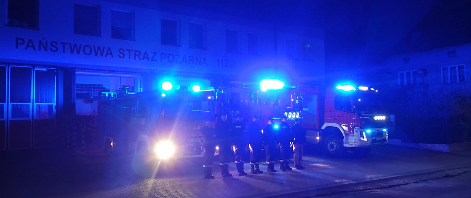 Na zdjęciu widać strażaków ustawionych w szeregu w pozycji na "baczność". Za nimi stoją pojazdy pożarnicze. W tle widać budynek strażnicy PSP.