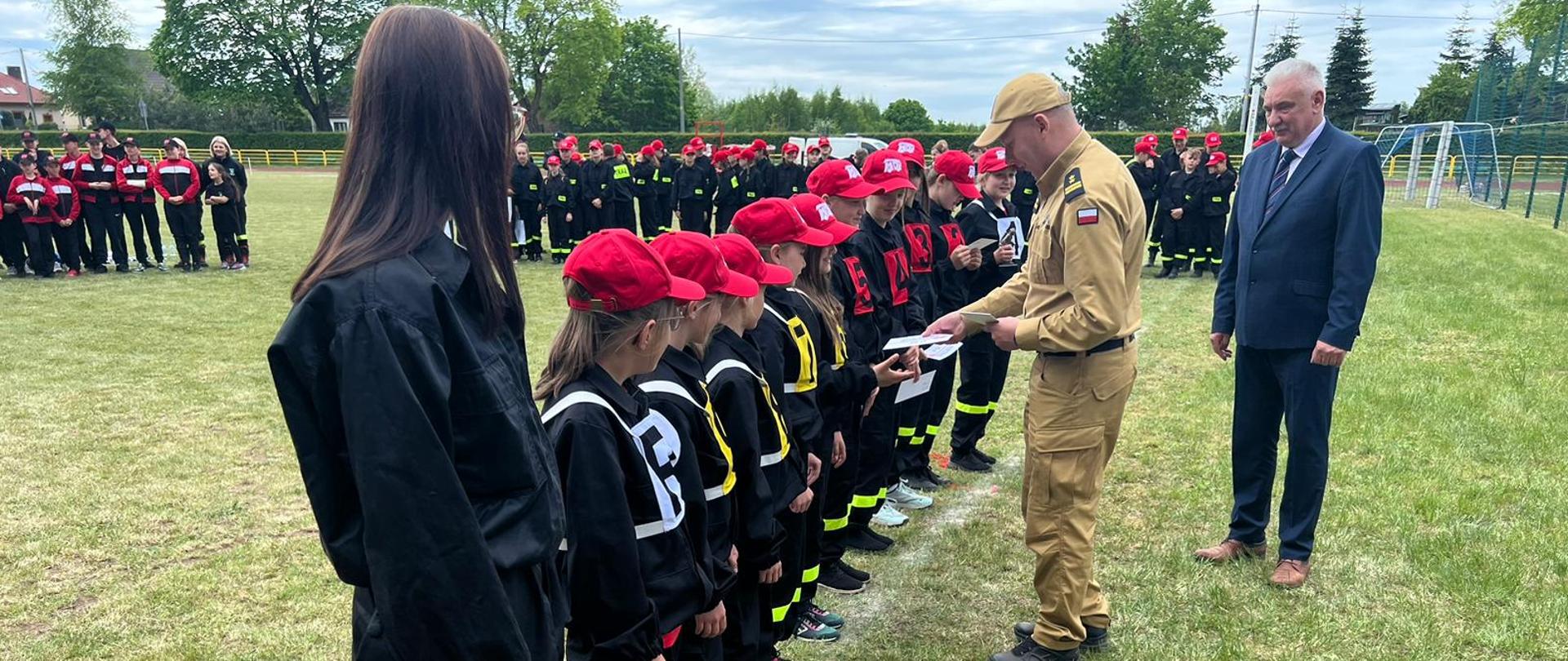 Komendant Powiatowy Państwowej Straży Pożarnej w Lęborku wraz z Wójtem Gminy Cewice wręczają nagrody jednej z drużyn dziewczęcych biorących udział w Powiatowych Zawodach Sportowo - Pożarniczych Młodzieżowych Drużyn Pożarniczych. Wszystkiemu przyglądają się z oddali trzy inne drużyny biorące udział w zawodach. 