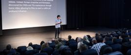 Na zdjęciu widoczna widownia w kinie, scena z osobą przemawiającą do mikrofonu, za przemawiającym widoczny jest ekran z treścią przemówienia.