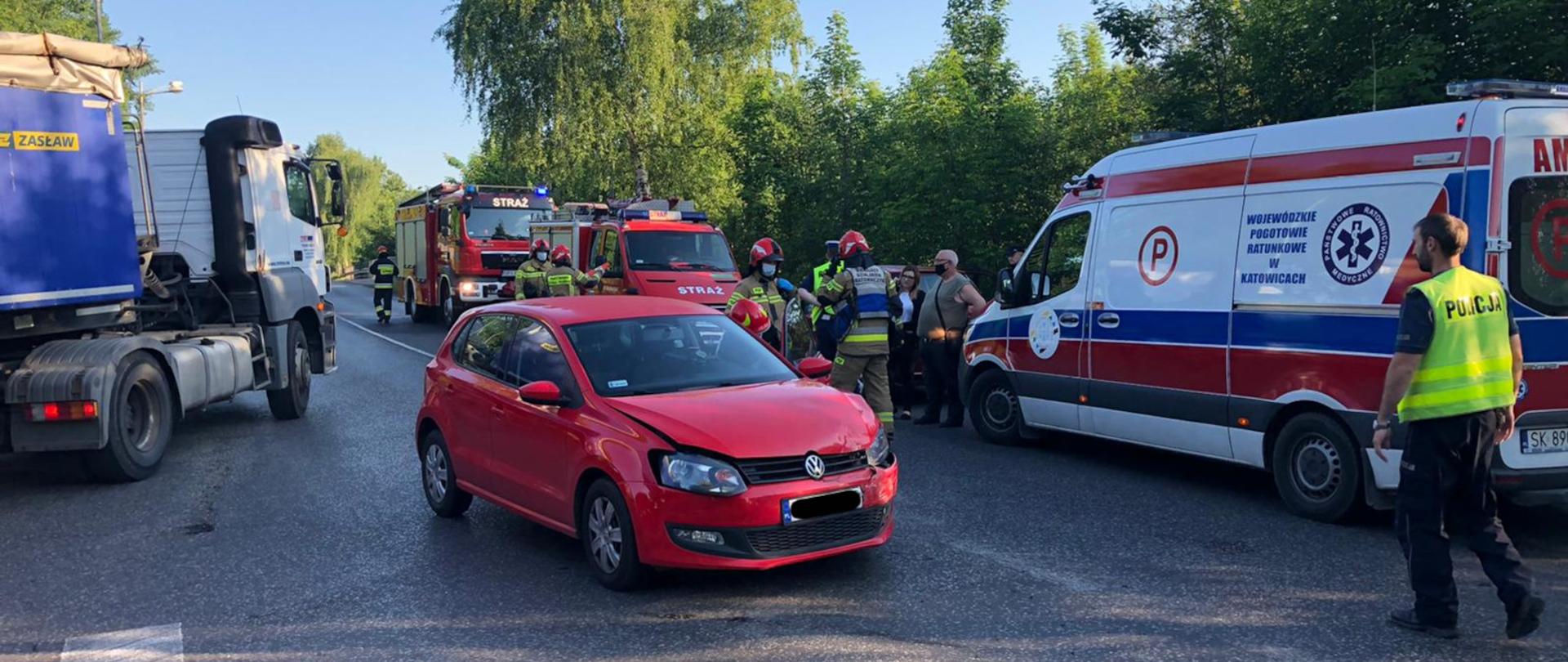 Zdjęcie przedstawia na pierwszym planie samochód uczestniczącym w kolizji. Z tyłu stoi karetka pogotowia, średni samochód ratowniczo - gaśniczy oraz lekki samochód ratownictwa technicznego. obok samochodów stoją policjanci i strażacy.