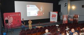 Szkolenie opiekunów MDP. Zdjęcie przedstawia salę kinową. Na tle ekranu, na którym wyświetlana jest prezentacja, przez mikrofon przemawia strażak w mundurze koloru piaskowego. Obok niego stoi koordynator projektu . PO obu stronach ekranu ustawione są roll-up oraz ścianka promocyjna. 