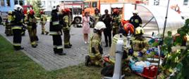 Strażacy w ubraniach specjalnych w hełmach strażackich zaopatrujący poszkodowane osoby ewakuowane ze szpitala w tle strażacy uczestniczący podczas ćwiczeń na szpitalu w Gorlicach w tm samochody pożarnicze i budynek szpitala.