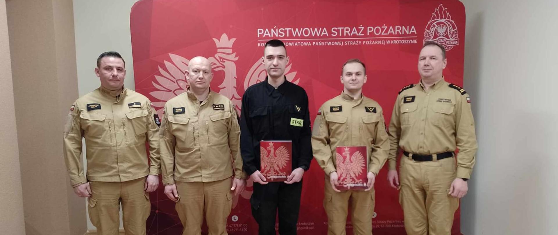 Zdjęcie grupowe przedstawia komendanta powiatowego PSP, dowództwo Jednostki Ratowniczo - Gaśniczej oraz awansowanych funkcjonariuszy na tle baneru komendy. 