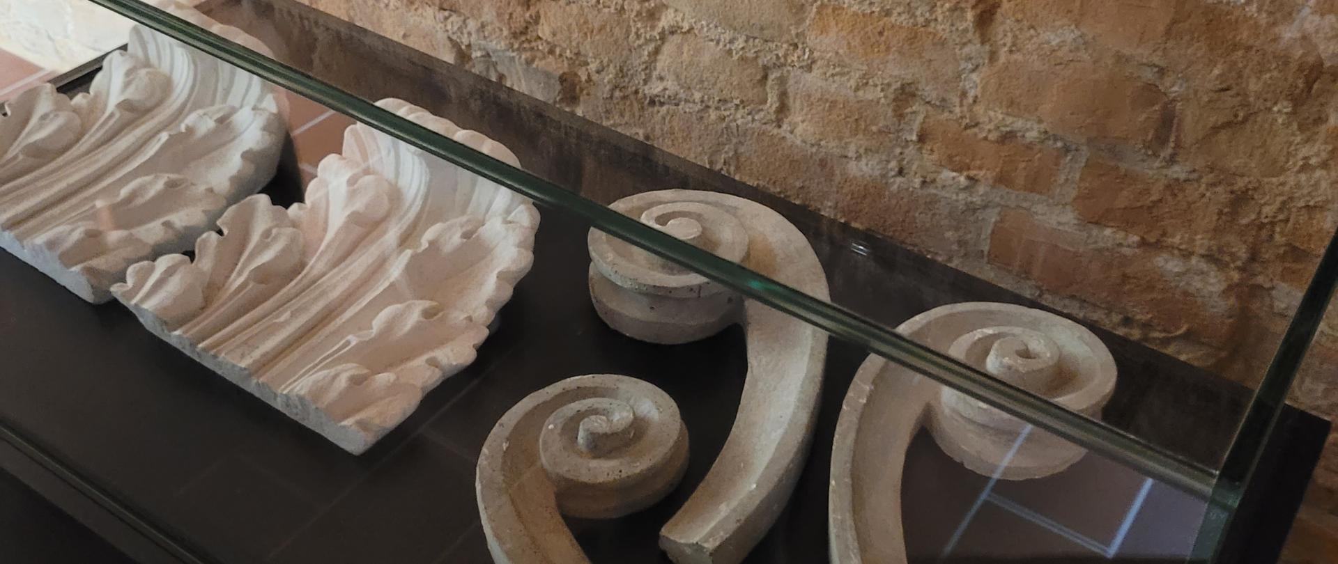 Na zdjęciu widać fragmenty kamiennych elementów dekoracyjnych, prawdopodobnie detali architektonicznych, umieszczone w przeszklonej gablocie na tle starej ceglanej ściany.