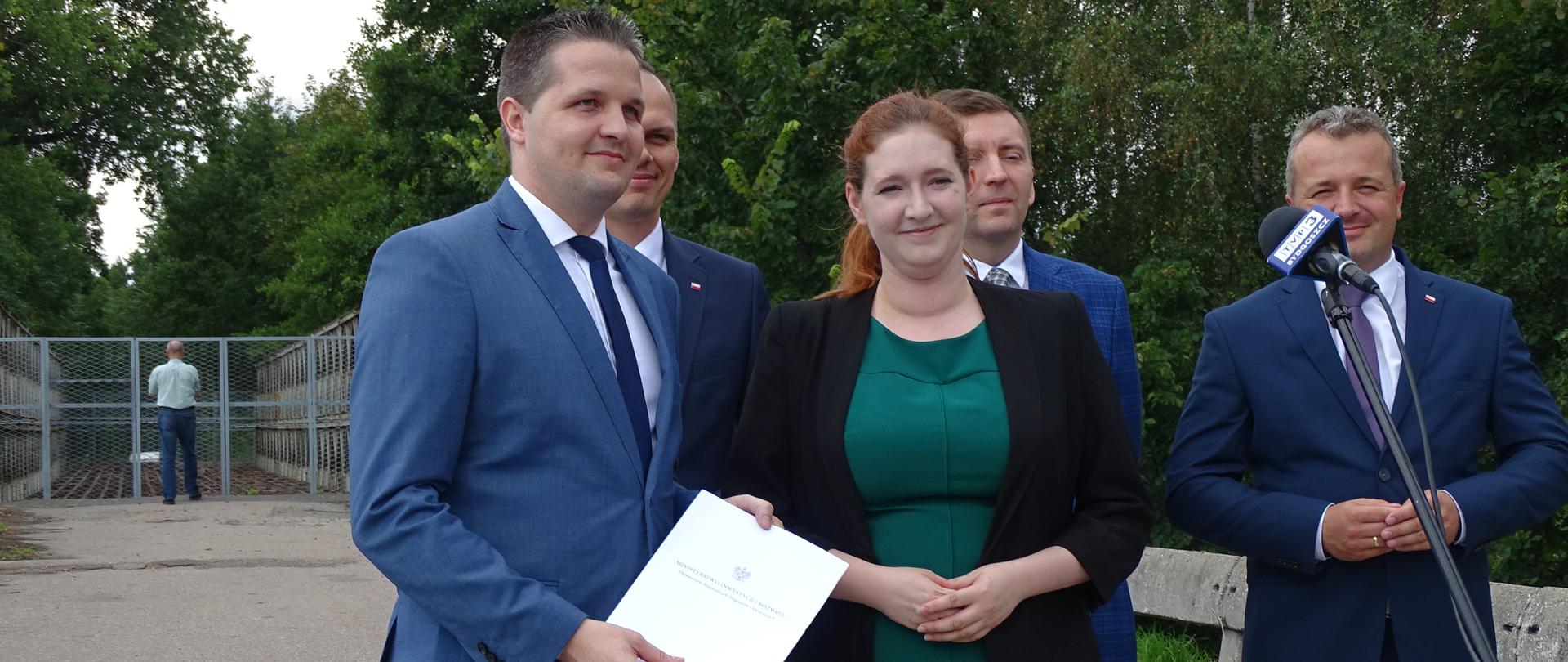 Zdjęcie w plenerze. Na drodze stoi grupa ludzi. Przed nimi mikrofon. Na pierwszym planie wiceminister Anna Gembicka.