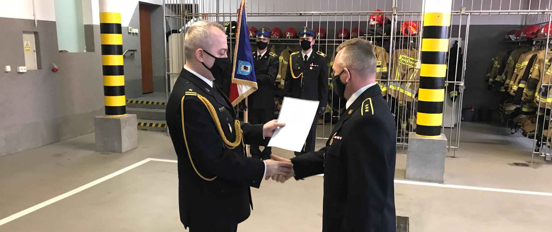mł. bryg. Paweł Gil, komendant powiatowy PSP w Kartuzach wręcza decyzje o zwolnieniu ze służby asp. sztab. Andrzejowi Brylowskiemu. W tle poczet sztandarowy.