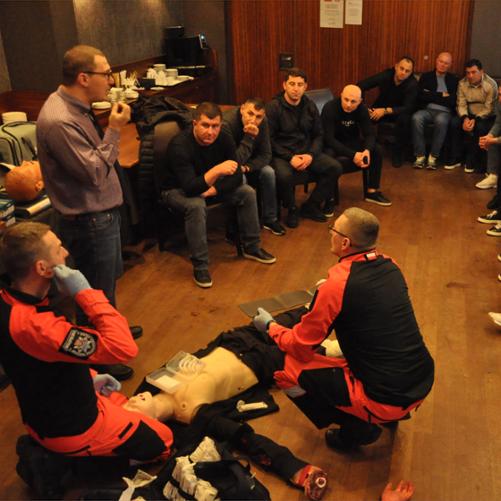 Grupa mężczyzn siedzi wokół leżącego na podłodze fantomu i słucha instruktora oraz dwóch ratowników demonstrujących udzielanie pierwszej pomocy