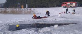 Na zdjęciu widoczne zamarznięty zalew w Dubiczach Cerkiewnych. w tle widoczny wóz strażacki, dwóch strażaków asekurujących sanie lodowe prowadzone przez strażaka. na saniach kolejny ratownik . widoczny poszkodowany w przeręblu.
