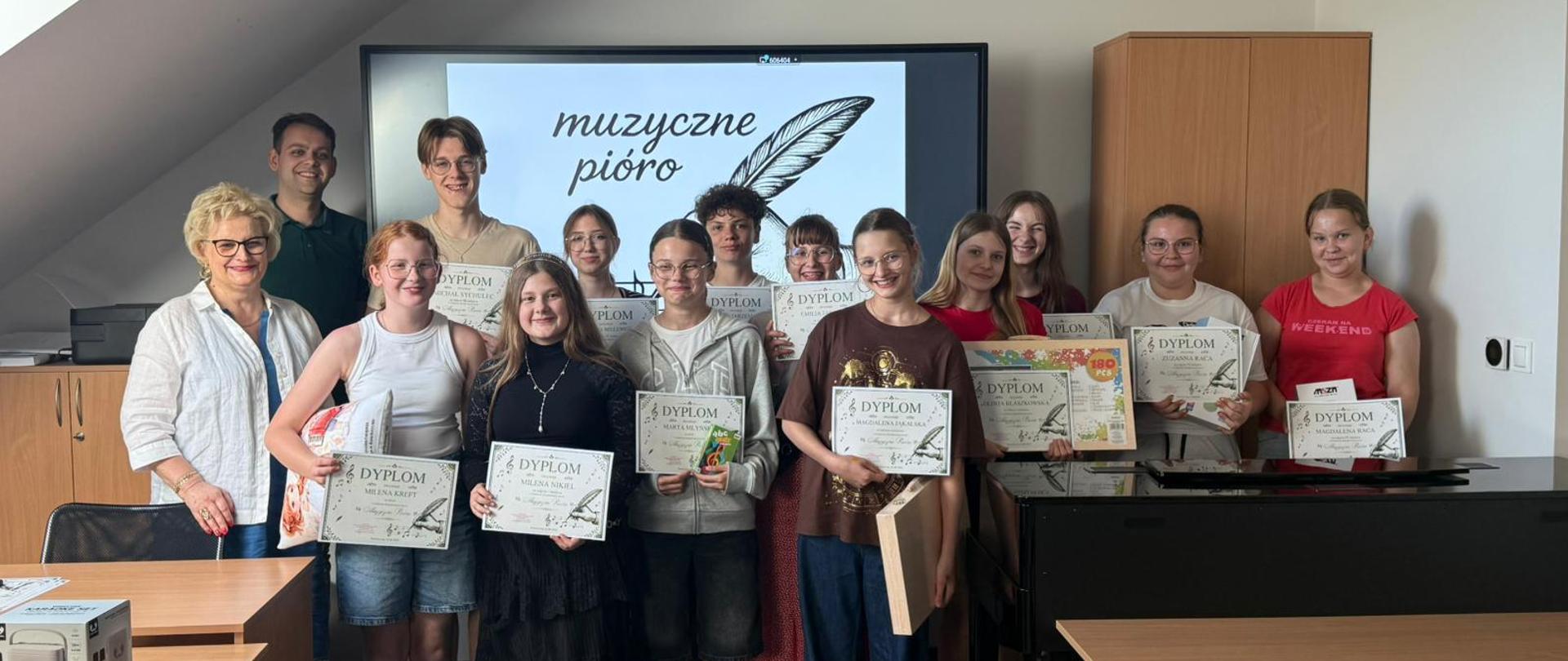 Grupa uczniów wraz z dwojgiem nauczycieli stoi w sali szkolnej podczas wręczenia dyplomów i nagród po szkolnym konkursie dyktand muzycznych „Muzyczne pióro”. Uczniowie trzymają w dłoniach dyplomy oraz upominki, a w tle na ekranie wyświetlony jest napis „muzyczne pióro” z grafiką pióra. Zdjęcie przedstawia uroczysty moment podsumowania konkursu i wspólne zdjęcie uczestników oraz organizatorów wydarzenia.