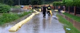 Na zdjęciu widać strażaków układających wał z worków z piaskiem. Wał ma chronić pobliskie zabudowania przed zalaniem. Na zdjęciu ulica zalana wodą.