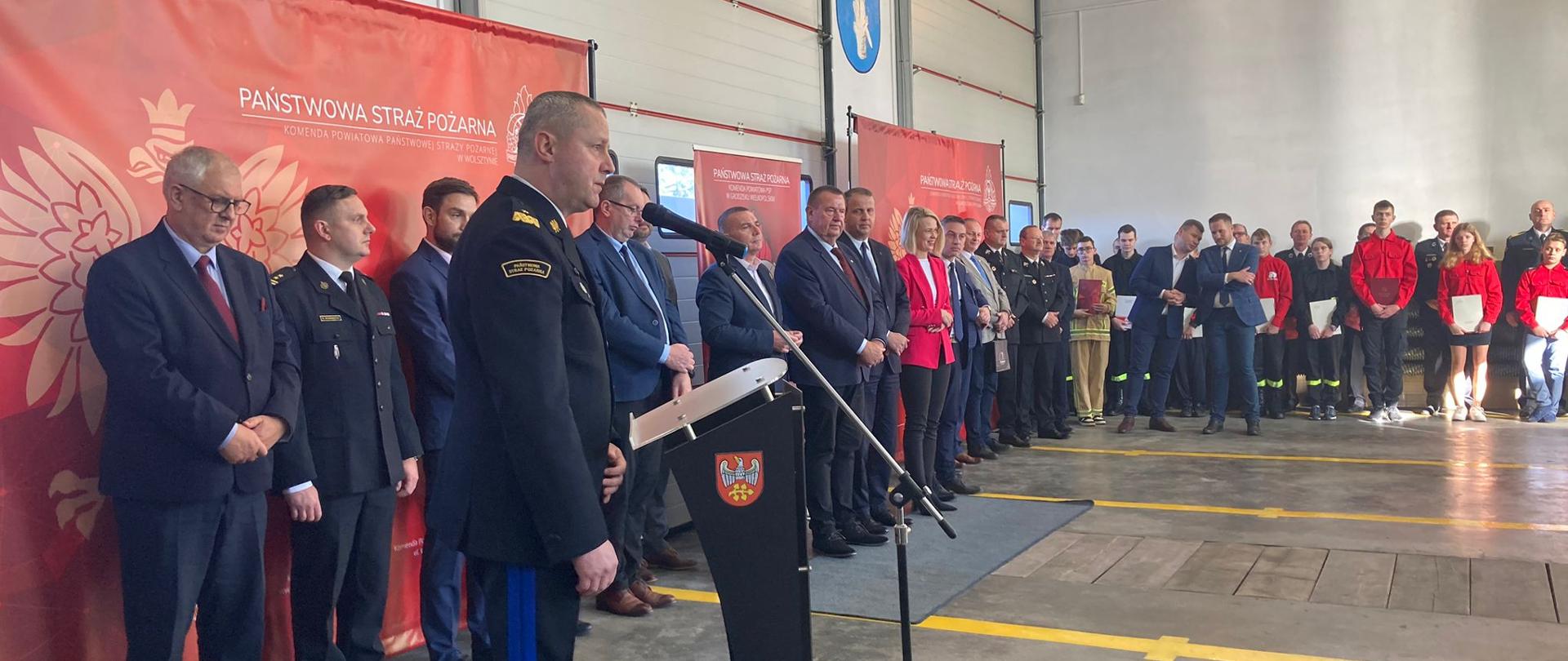 Na pierwszym planie znajduje się Wielkopolski Komendant Wojewódzki PSP przemawiający do uczestników uroczystości. Na drugim planie stoją oficjele oraz zaproszeni goście.