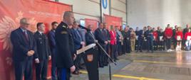 Na pierwszym planie znajduje się Wielkopolski Komendant Wojewódzki PSP przemawiający do uczestników uroczystości. Na drugim planie stoją oficjele oraz zaproszeni goście.