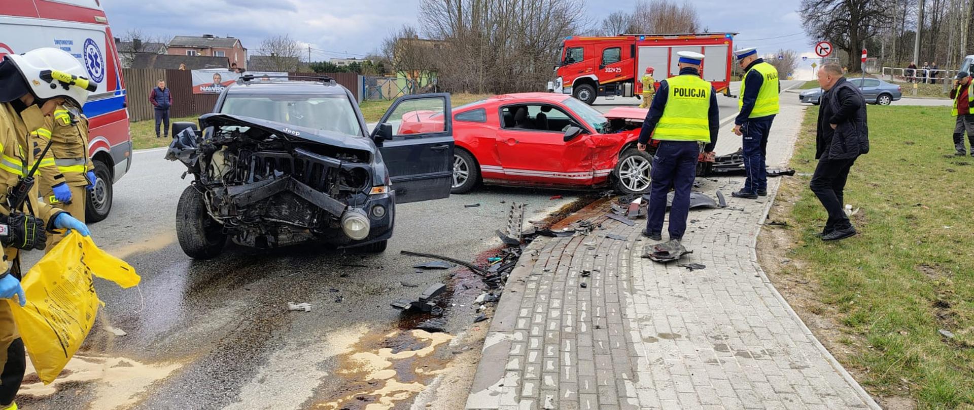 Zdjęcie przedstawia miejsce wypadku: w centralnym punkcie oba zniszczone pojazdy, z prawej strony stoją funkcjonariusze Policji a z lewej widać ambulans i strażaków usuwających wycieki z jezdni.