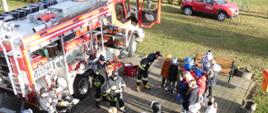 Zdjęcie wykonane jest na świeżym powietrzu i przedstawia strażaków OSP, którzy prezentują dzieciom swój samochód pożarniczy. W samochodzie pootwierane są skrytki i pokazany jest sprzęt będący na jego wyposażeniu. Strażacy są w ubraniach specjalnych, na głowach mają czapki. W tle zdjęcia widać samochód operacyjny PSP. 