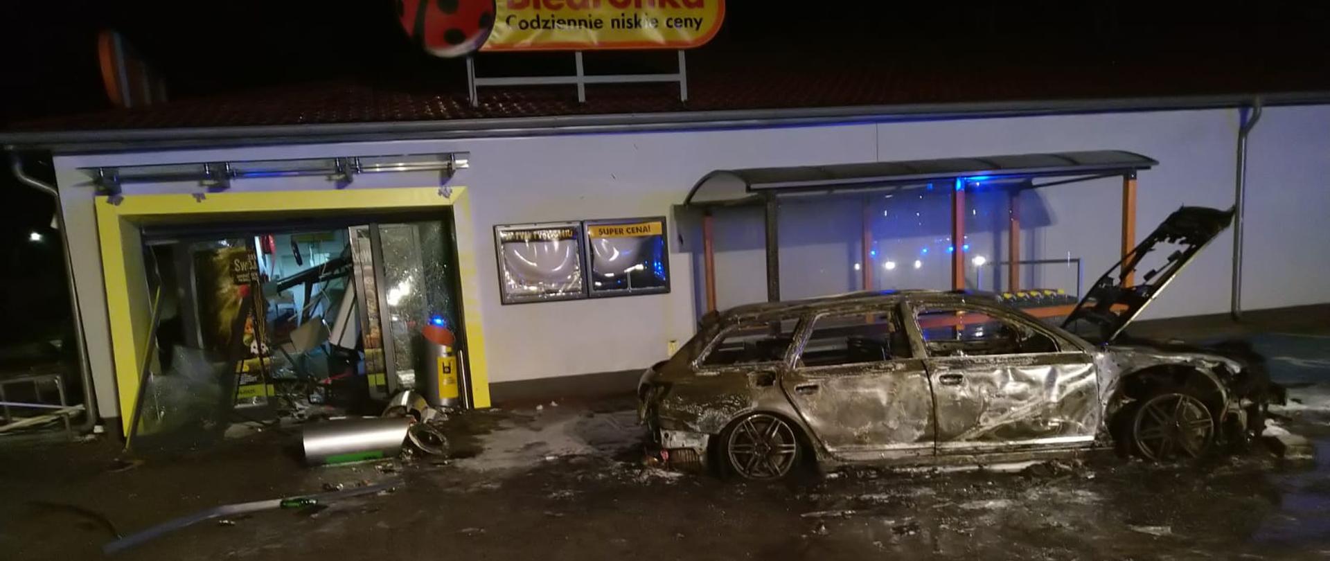 Zdjęcie przedstawia spalony wrak samochodu osobowego, a obok wejście do Biedronki, w którym został wysadzony bankomat.