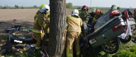 Tragiczny wypadek. Droga z Sarnówki do Łaszczyna. Obok drzewa znajduje sie wrak samochodu osobowego, który brał udział w wypadku. Jest koloru srebrnego. Wokół auta strażacy prowadzący akcje ratowniczą. Jeden z nich pracuje nozycami hydraulicznymi.W tle pola, turbiny wiatrowe i zabudowania.