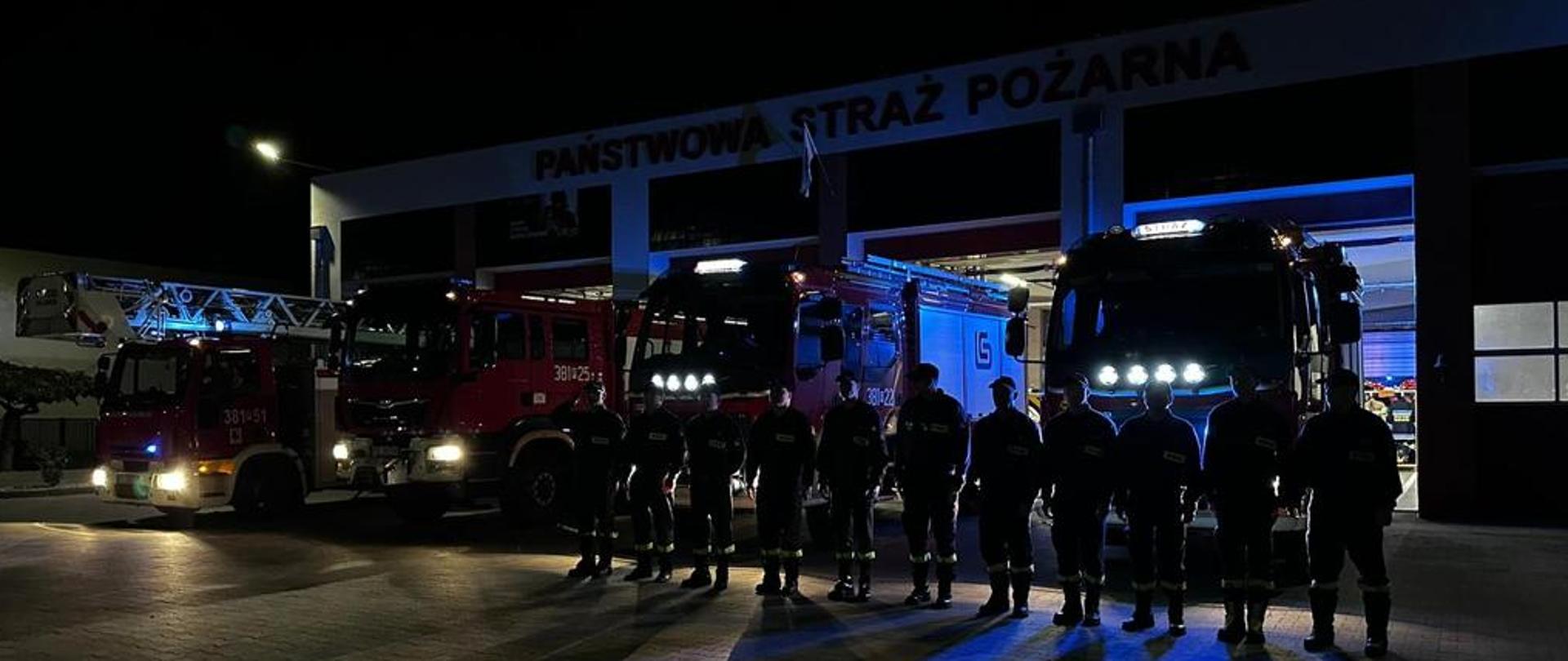 Strażacy stoją w szeregu na tle samochodów strażackich.