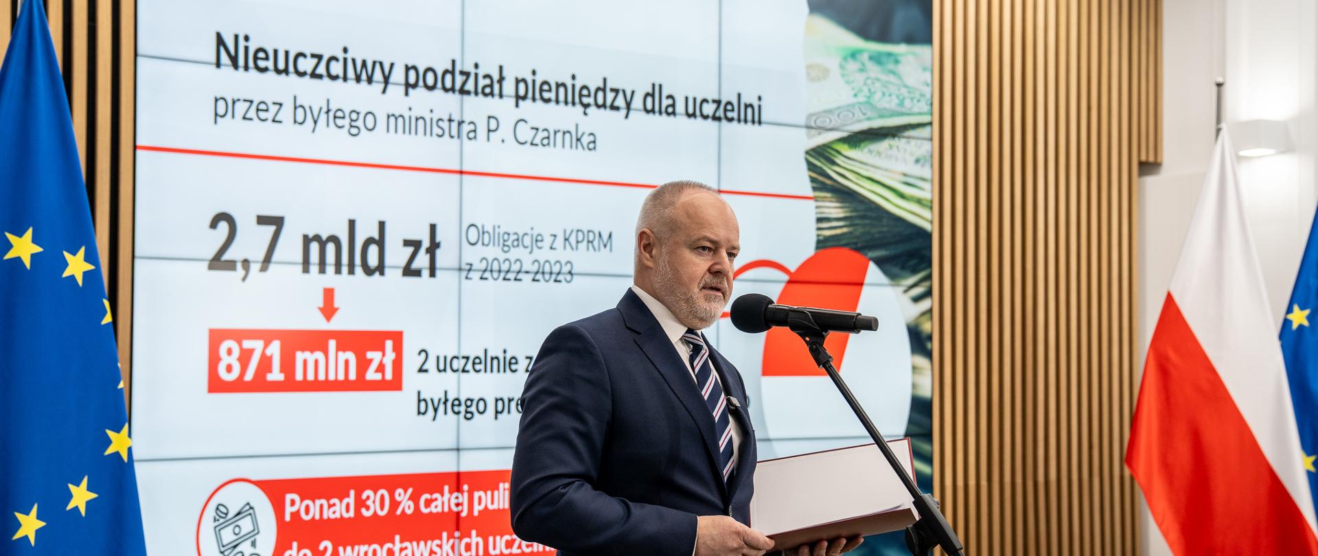 Wiceminister Gzik stoi na sali i mówi do mikrofonu na stojaku, za nim pod ścianą z drewnianych listewek flagi Polski i UE, na wbudowanym w ścianę ekranie napis Nieuczciwy podział pieniędzy dla uczelni.