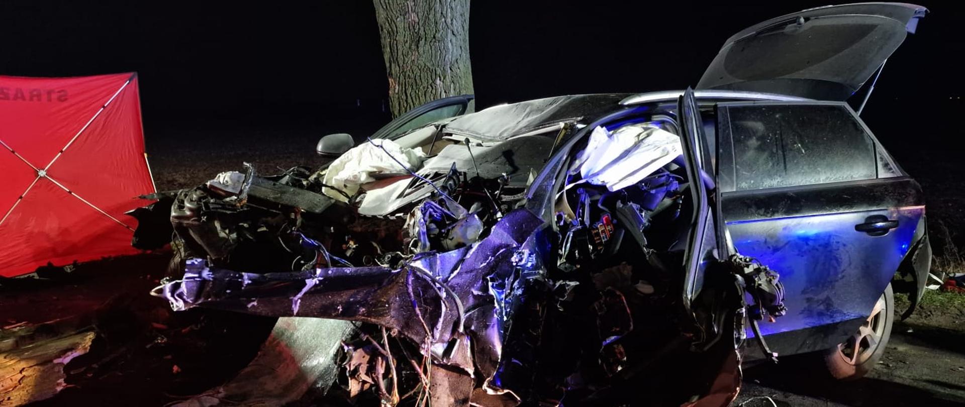 Widać mocno uszkodzony samochód od przodu i lewego boku, za nim jest drzewo, jest ciemno