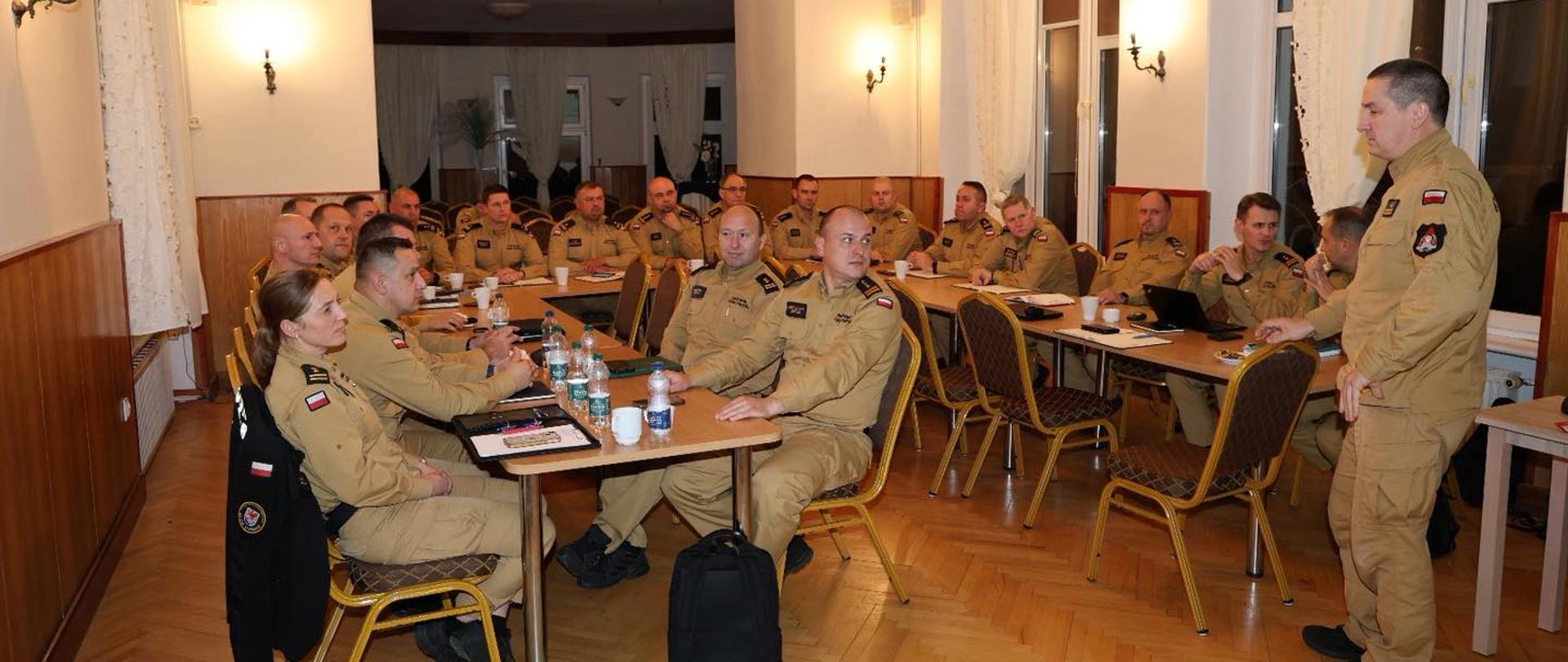 Na zdjęciu widać grupę osób w jednakowych mundurach koloru piaskowego, siedzących przy stołach w sali o drewnianej podłodze i jasnych ścianach. Wszyscy są zwróceni w stronę mężczyzny, który stoi po prawej stronie zdjęcia i najwyraźniej prowadzi spotkanie lub szkolenie. Na stołach stoją butelki z wodą, kubki i notatniki. Pomieszczenie jest dobrze oświetlone, ma kilka dużych okien z białymi firanami i żyrandole z żarówkami w stylu klasycznym na suficie z drewnianą kratownicą. Wszyscy uczestnicy są skupieni i uważnie słuchają prowadzącego.