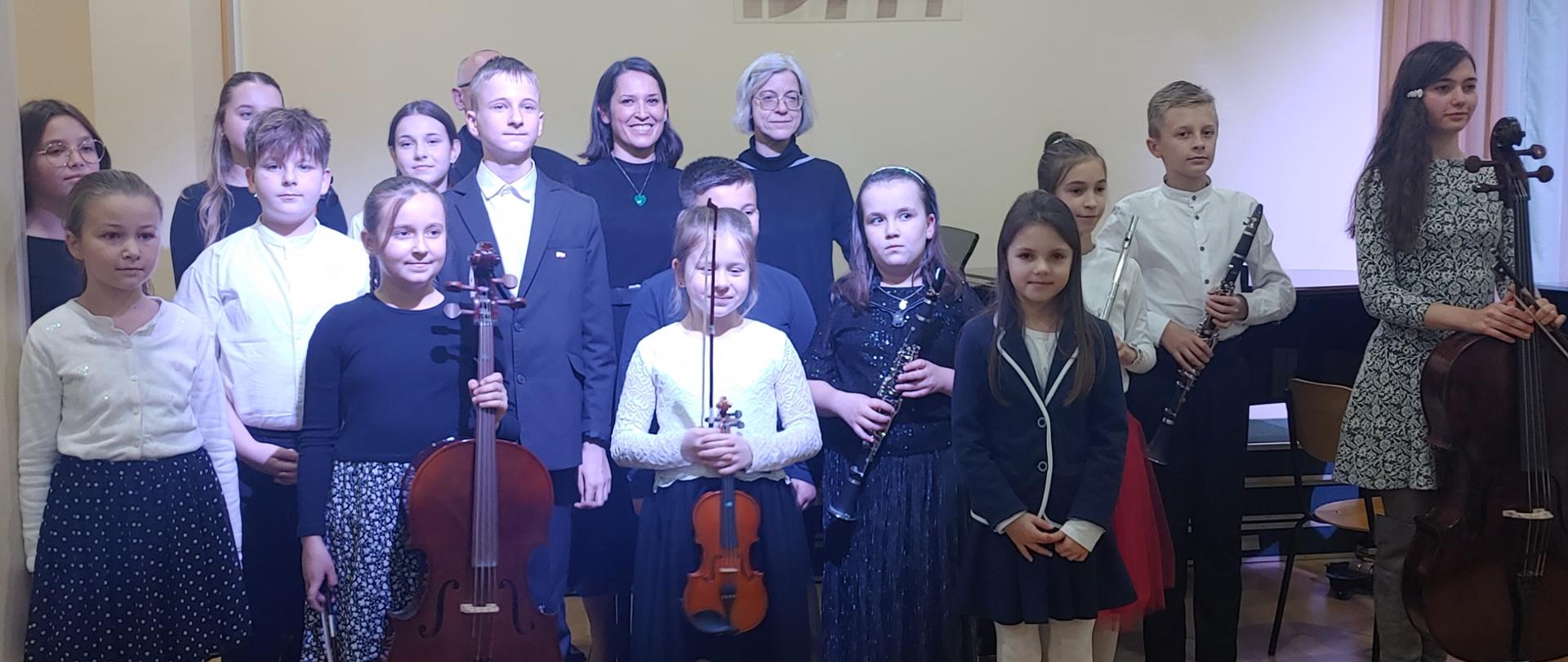 Na zdjęciu widać uczniów i nauczycieli w strojach koncertowych pozujących do zdjęcia grupowego na tle żółtej ściany. Niektórzy uczniowie trzymają instrumenty wiolonczele , klarnety, flet i skrzypce. Na ścianie wisi logo szkoły PSM . Uczestnicy koncertu stoją na parkiecie na scenie w sali koncertowej.