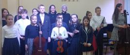 Na zdjęciu widać uczniów i nauczycieli w strojach koncertowych pozujących do zdjęcia grupowego na tle żółtej ściany. Niektórzy uczniowie trzymają instrumenty wiolonczele , klarnety, flet i skrzypce.