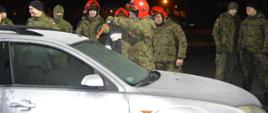 Współdziałanie z WOT. Plac Komendy Powiatowej PSP w Rawiczu. Żołnierze i strażacy stoją wokół samochodu osobowego będącego obiektem ćwiczeń. Jeden z nich przy pomocy specjalnego narzędzia - piły tnie przednią szybę auta. Czterech żołnierzy ma na głowach czerwone hełmy, pozostali są w czapkach zimowych.